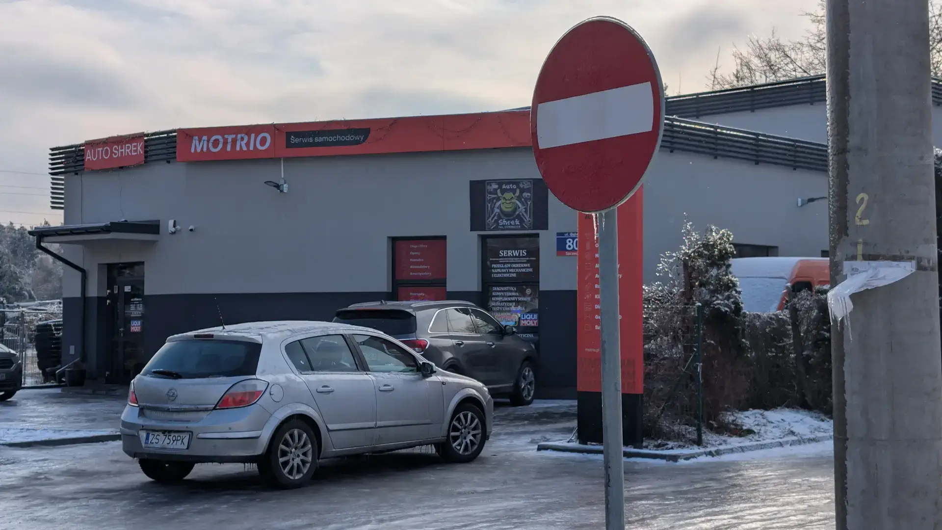 Oznakowany budynek serwisu partnerskiego Motrio Autoshrek oraz zaparkowany srebrny Opel Astra przed naprawą.