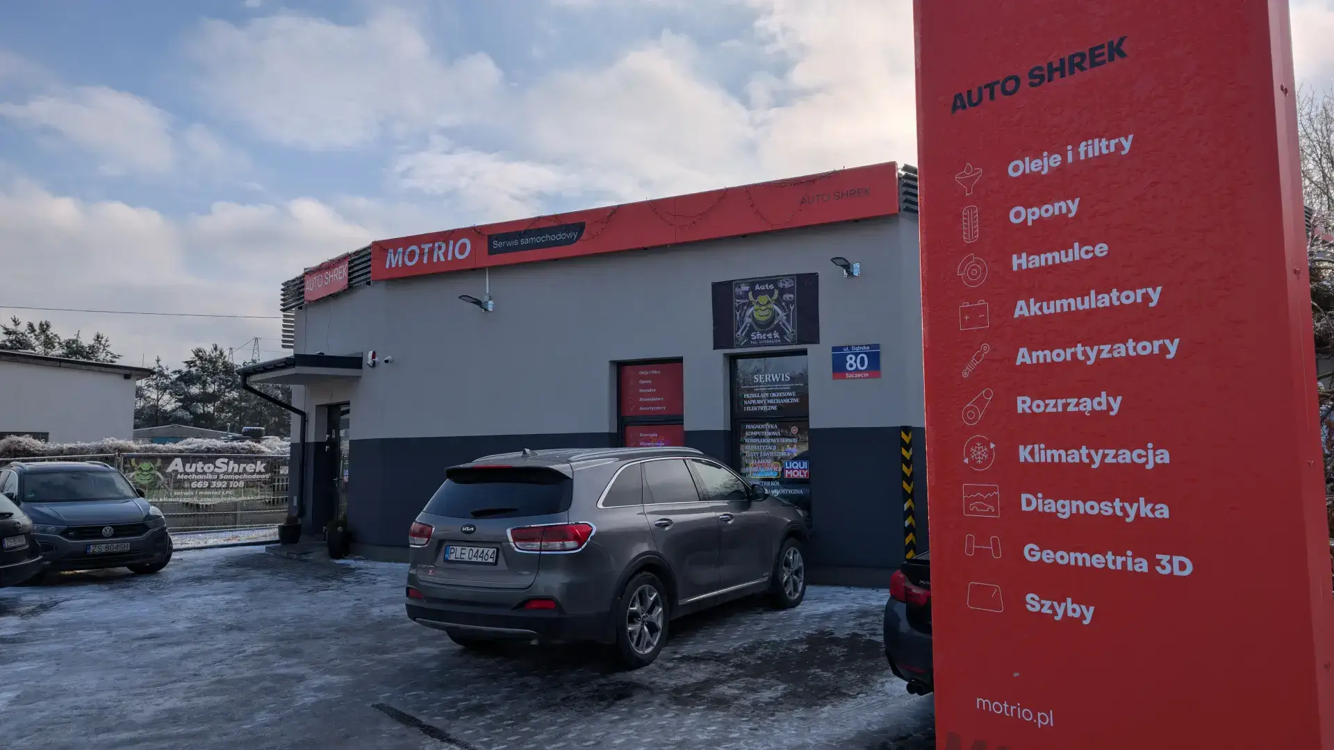 Czerwony pylon reklamowy przy ulicy wymieniający usługi: oleje, opony, hamulce, rozrządy i serwis szyb.