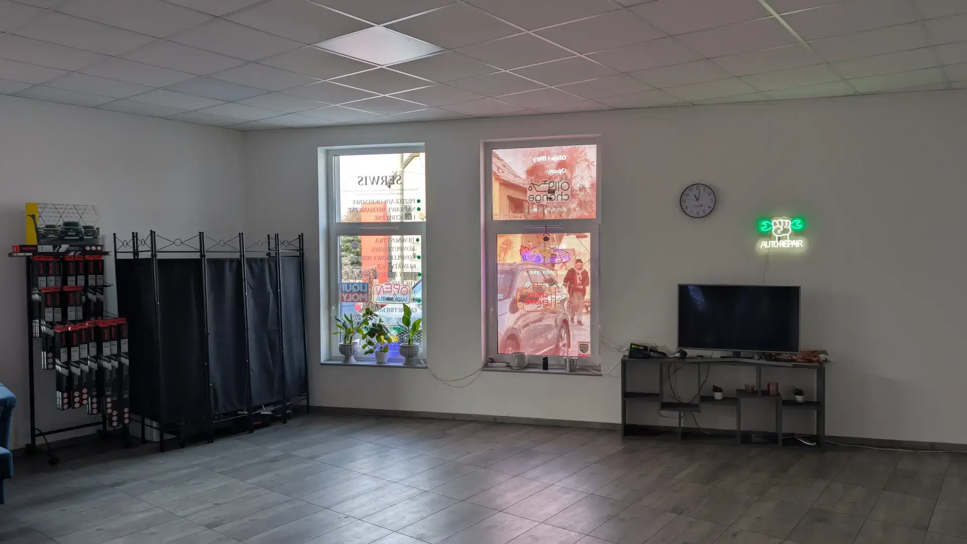 Przestronna poczekalnia z telewizorem, neonem "Auto Repair" i stojakiem z wycieraczkami samochodowymi.